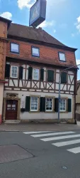 Ein historisches Fachwerkhaus mit einem roten Ziegeldach und grünen Fensterläden. Es steht an einer Kreuzung mit einem Zebrastreifen.