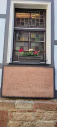 Ein Fenster mit bunten Blumenkästen und einem Gitter. Darunter befindet sich eine Tafel mit historischer Inschrift.