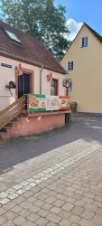 Ein kleines Restaurant mit einer bunten Werbetafel steht an einer gepflasterten Straße. Im Hintergrund sind Wohngebäude und ein blauer Himmel zu sehen.