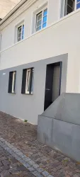 Ein modernes Gebäude mit einer grauen Wand und großen Fenstern. Der Eingang ist durch eine schwarze Tür gekennzeichnet.