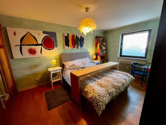 Ein gemütliches Schlafzimmer mit einem großen Bett und bunten Kunstwerken an der Wand. Das Zimmer hat ein Fenster mit natürlichem Licht und eine angenehme Atmosphäre.