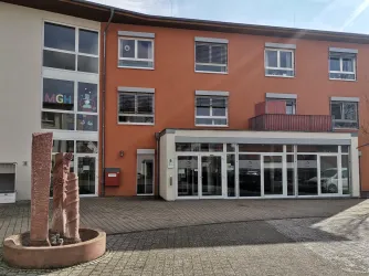 Ein modernes Gebäude mit orangefarbener Fassade und großen Fenstern. Vor dem Haus steht eine Skulptur.