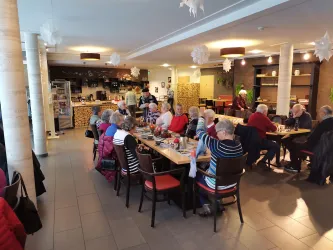 Eine gemütliche Cafeteria mit vielen älteren Menschen, die an Tischen sitzen und sich unterhalten. Im Hintergrund ist eine kleine Theke und ein freundliches Ambiente zu sehen.