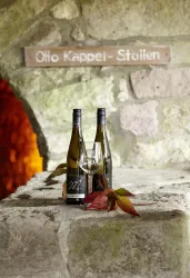 Zwei Weinflaschen und ein Weinglas stehen auf einem steinernen Tisch. Im Hintergrund ist ein Schild mit dem Namen „Otto Kappel-Stollen“ zu sehen.