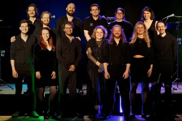 Eine Gruppe von Menschen in schwarzer Kleidung steht zusammen auf einer Bühne. Sie lächeln und posieren freundlich für das Foto.