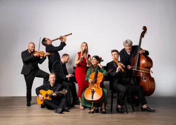 Eine Gruppe von Musikern steht zusammen, die verschiedene Instrumente spielen. Die Musiker sind in festlicher Kleidung und zeigen eine freundliche Atmosphäre.