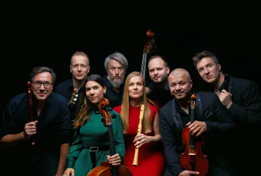 Eine Gruppe von acht Musikern steht in einem dunklen Hintergrund. Sie halten verschiedene Musikinstrumente und lächeln in die Kamera.