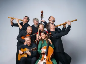 Eine Gruppe von Musikern mit verschiedenen Instrumenten steht zusammen. Sie lächeln und posieren fröhlich in einem neutralen Hintergrund.