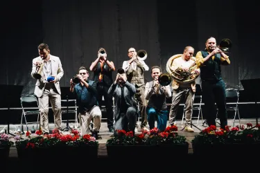 Eine Gruppe von Musikern steht auf der Bühne und spielt verschiedene Blasinstrumente. Im Vordergrund sind bunte Blumenarrangements zu sehen.