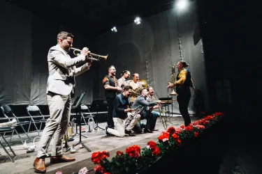 Eine lebhafte Musikszene mit mehreren Musikern auf einer Bühne. Ein Trompeter spielt, während andere Instrumente und Gesang zu hören sind.