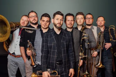 Eine Gruppe von acht Musikern steht zusammen und hält verschiedene Blechblasinstrumente. Sie tragen elegante Kleidung und lächeln in die Kamera.