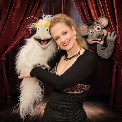 Eine lachende Frau trägt zwei Puppen, eine weiße und eine graue, in den Händen. Der Hintergrund ist mit roten Vorhängen dekoriert.