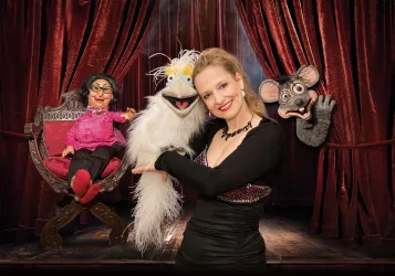 Eine Frau steht auf der Bühne und hält eine Puppenfigur. Im Hintergrund sind zwei weitere Puppen in einem Bühnenarrangement zu sehen.