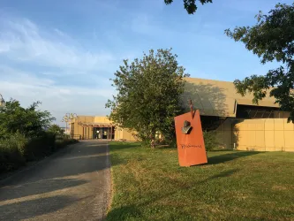 Ein moderner Gebäude mit einer klaren Fassade und einem großen Schild im Vordergrund. Der Weg ist von Bäumen umgeben und das Licht ist sanft.