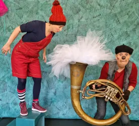Zwei Darsteller in bunten Kostümen stehen auf einer Bühne. Einer schaut neugierig auf ein großes Holzblasinstrument, während der andere es hält.