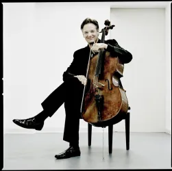 Ein Musiker sitzt entspannt auf einem Stuhl und hält ein Cello. Die Aufnahme hat einen modernen, klaren Hintergrund.