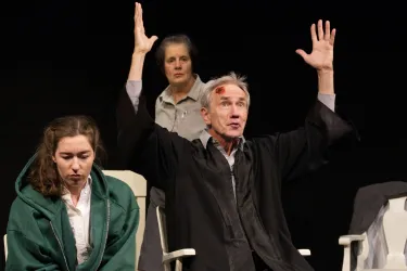 Eine dramatische Szene auf der Bühne mit drei Schauspielern. Der Mann in schwarzer Kleidung hebt die Hände, während die beiden Frauen aufmerksam zuschauen.