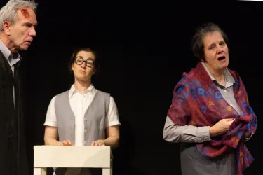 Drei Schauspieler auf einer Bühne in einem Theater. Sie zeigen intensive Emotionen und interagieren miteinander.