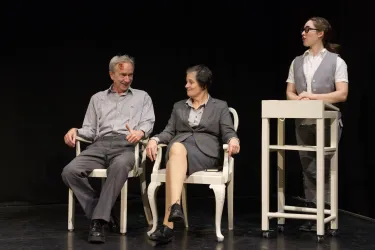Drei Personen auf einer Bühne in einem Theater. Zwei Personen sitzen auf Stühlen, während eine dritte Person an einem Tisch steht.