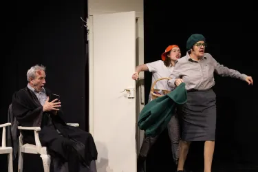 Eine Theaterszene mit drei Personen. Eine Frau in einem Anzug und mit einer Mütze verlässt energisch den Raum, während ein Mann im Hintergrund sitzt.