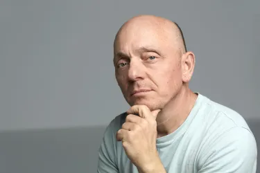 Ein nachdenklicher Mann mit Glatze trägt ein hellblaues T-Shirt. Er hat die Hand am Kinn und blickt direkt in die Kamera.