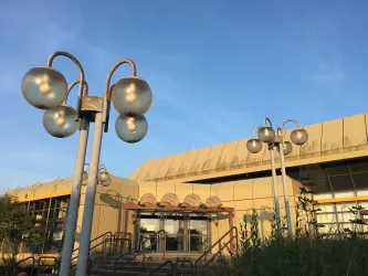 Ein modernes Gebäude mit einer gelben Fassade und verschiedenen Lampen im Vordergrund. Der Himmel ist klar und blau.