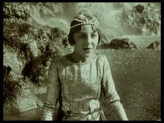 Ein Mädchen in einem glitzernden, eleganten Kleid sitzt am Wasserfall. Sie hat eine Haarband und schaut überrascht in die Kamera.