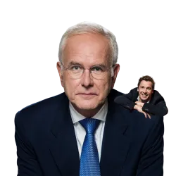 Ein älterer Mann in einem dunklen Anzug mit Brille schaut direkt in die Kamera. Im Hintergrund ist ein jüngerer Mann zu sehen, der entspannt wirkt.