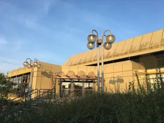 Ein modernes Gebäude mit gelber Fassade und runden Fenstern. Im Vordergrund sind Lampen und viel Grün zu sehen.