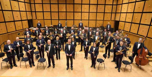 Ein Orchester steht auf der Bühne in eleganter Kleidung. Es besteht aus Musikern, die verschiedene Instrumente spielen und lächeln.