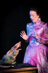 Eine Frau in einem eleganten lila Kleid steht überrascht vor einem Puppen-Wolf. Der Wolf hat einen merkwürdigen Ausdruck und scheint zu sprechen.