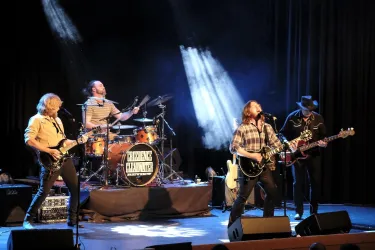 Eine Band spielt auf der Bühne vor einem dunklen Hintergrund. Die Musiker sind leidenschaftlich bei ihrem Auftritt und nutzen verschiedene Instrumente.
