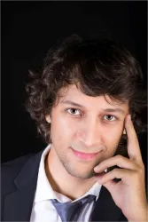 Ein junger Mann mit lockigem Haar, der in die Kamera lächelt. Er trägt einen Anzug und hält eine Hand an sein Gesicht.