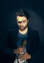 Ein Mann mit einem Mikrofon in der Hand und einem Klebezettel mit der Aufschrift "POP" auf der Stirn. Er trägt ein lässiges Outfit und schaut direkt in die Kamera.