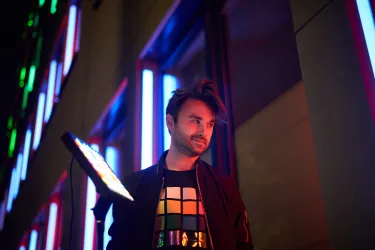 Ein Mann steht nachts vor einem Gebäude mit bunten Lichtern. Er trägt ein T-Shirt mit einem geometrischen Muster und hält eine Leuchte in der Hand.