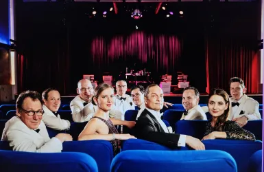Eine Gruppe von Menschen in eleganter Kleidung sitzt in blauen Stühlen. Im Hintergrund ist eine Bühne mit Lichtern und Musikinstrumenten zu sehen.