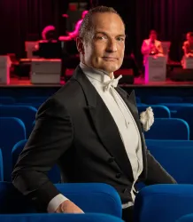 Ein eleganter Mann in einem Smoking sitzt in einem blauen Sessel. Im Hintergrund sind Bühnenlichter und Musiker zu sehen.