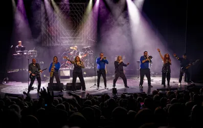 Eine Live-Band tritt auf einer Bühne auf, umgeben von Lichtern und Rauch. Die Musiker und Sänger unterhalten das Publikum mit energiegeladener Musik.