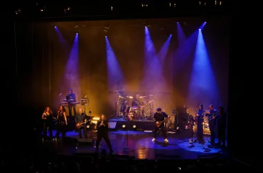 Ein Live-Konzert auf einer Bühne mit mehreren Musikern und Sängern. Bunte Lichter und ein begeistertes Publikum schaffen eine energische Atmosphäre.