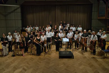 Eine große Gruppe von Menschen steht lächelnd auf einer Bühne. Sie sind Musiker in Konzertkleidung und halten verschiedene Musikinstrumente.