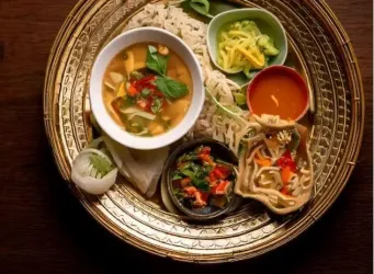 Ein traditionelles Gericht auf einem goldenen Teller mit einer Schüssel Suppe, Reisnudeln und verschiedenen Beilagen. Frische Kräuter und Gemüse ergänzen die appetitliche Präsentation.