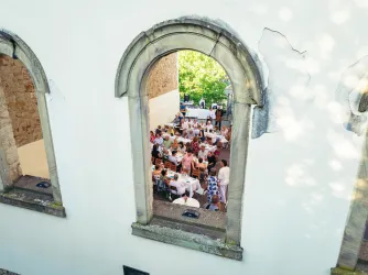 Ein festlich gedeckter Tisch mit vielen Gästen, die draußen speisen. Der Blick erfolgt durch ein großes Fenster in einer historischen Mauer.
