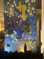Eine farbenfrohe Wandmalerei mit Engeln und anderen Figuren vor einem nächtlichen Himmel. Im Vordergrund sind Silhouetten von Stadtgebäuden und Sternen zu sehen.