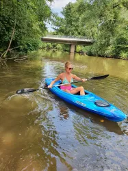 Eine Frau paddelt in einem blauen Kajak auf einem ruhigen Fluss. Im Hintergrund ist eine Brücke und üppige Bäume zu sehen.
