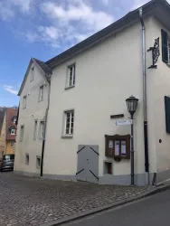 Ein helles, zweigeschossiges Gebäude an einer Straßenecke. Es gibt Kaffeehäuser und eine Straßenlaterne in der Nähe.
