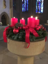 Ein Adventskranz mit vier roten Kerzen steht auf einem steinernen Podest. Er ist mit Weihnachtskugeln und roten Schleifen dekoriert.