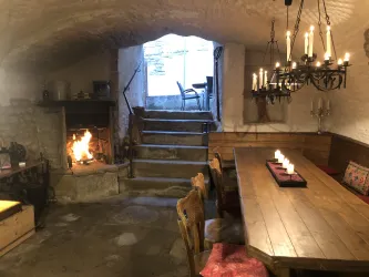Ein gemütlicher Raum mit einem großen Holztisch und einer Feuerstelle. Im Hintergrund sind Treppen und ein Fenster, das Licht hereinlässt.