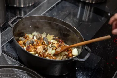 Ein großer Kochtopf mit gebratenem Gemüse und Fleisch steht auf einem modernen Kochfeld. Dampf steigt auf, während jemand mit einem Holzlöffel umrührt.