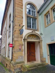 Ein historisches Gebäude mit einer schönen Eingangstür und großen Fenstern. Nebenan steht ein modernes Gebäude in sanften Farben.