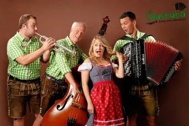 Eine Gruppe von Musikern in traditioneller Tracht spielt Instrumente. Eine Frau in einem Dirndl singt fröhlich mit drei männlichen Musikern.